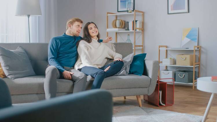 Paar sitzt auf grauer Couch in modernem Umfeld und schaut gemeinsam in eine Richtung, wahrscheinlich Richtung Fernseher