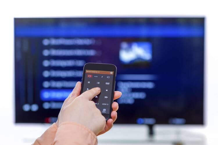 Hand hält Smartphone mit Fernbedienungs Applikation in Richtung eines Fernsehers