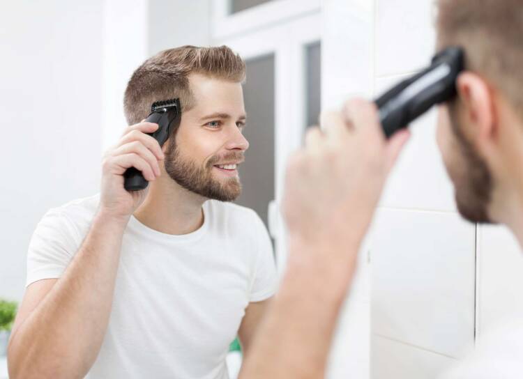 Glücklich schauender junger Mann schneidet sich die Haare mit einem Haarschneider