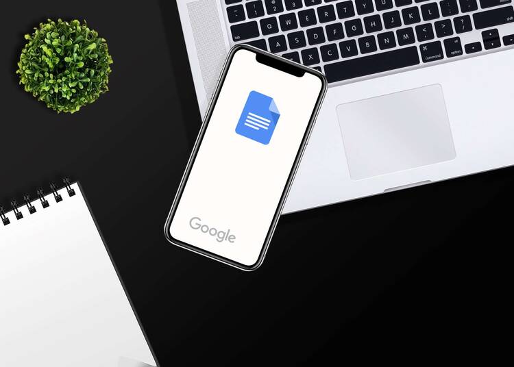 Smartphone mit Google Docs Logo liegt auf Macbook in Arbeitsumgebung