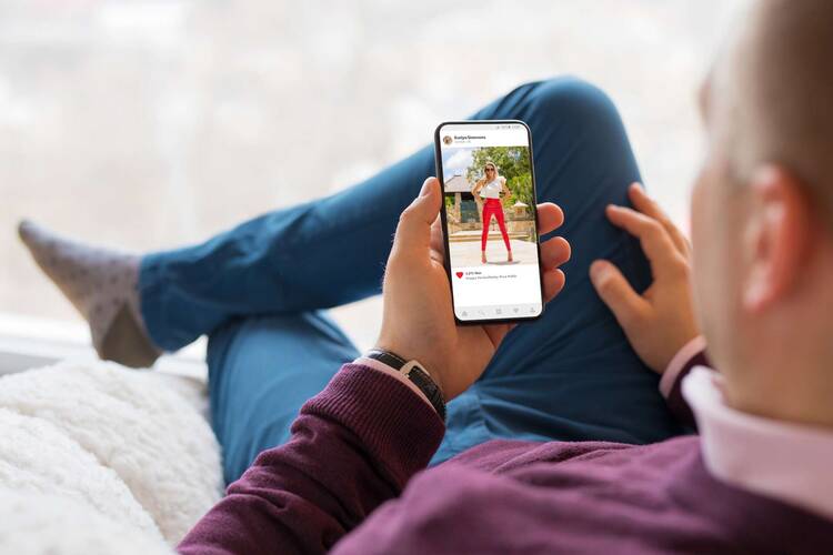 Mann sitzt entspannt in Socken und Jeans und hält dabei Smartphone mit offener Instagram Applikation in der Hand