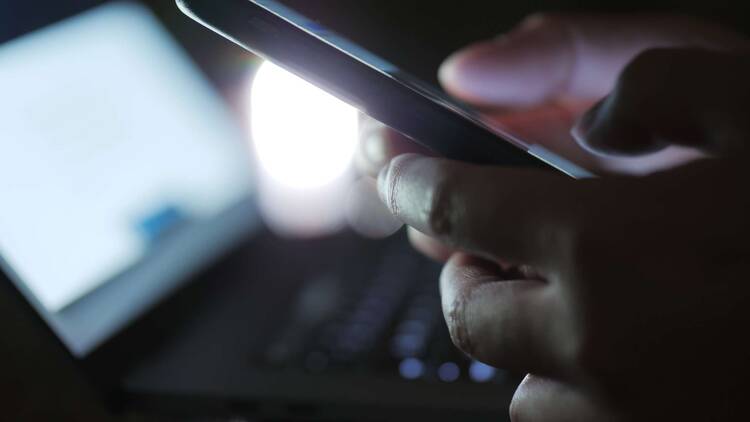 Finger tippen auf Smartphone, dunkle Umgebung, Laptop im Hintergrund
