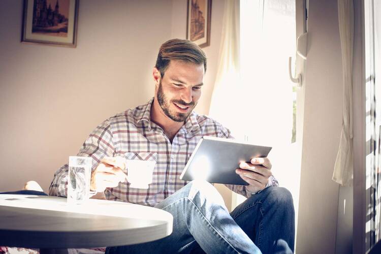 Mann mit Kaffeetasse im Karohemd sitzt an rundem Tisch mit iPad in der Hand