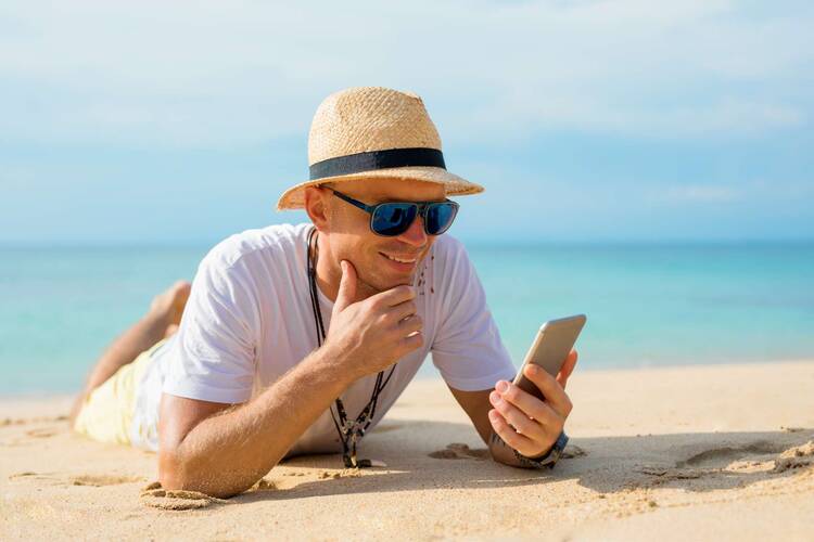 Mann mit Hut liegt mit Handy in der Hand am Strand