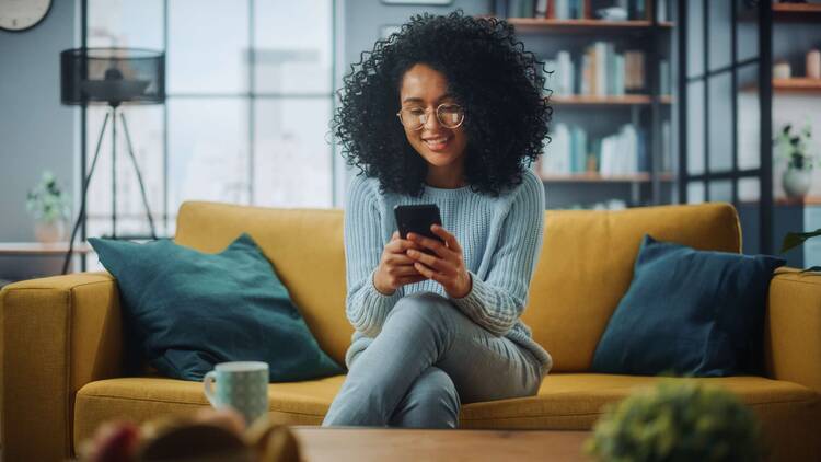 Frau sitzt lächelnd auf gelbem Sofa und schaut auf ihr Smartphone