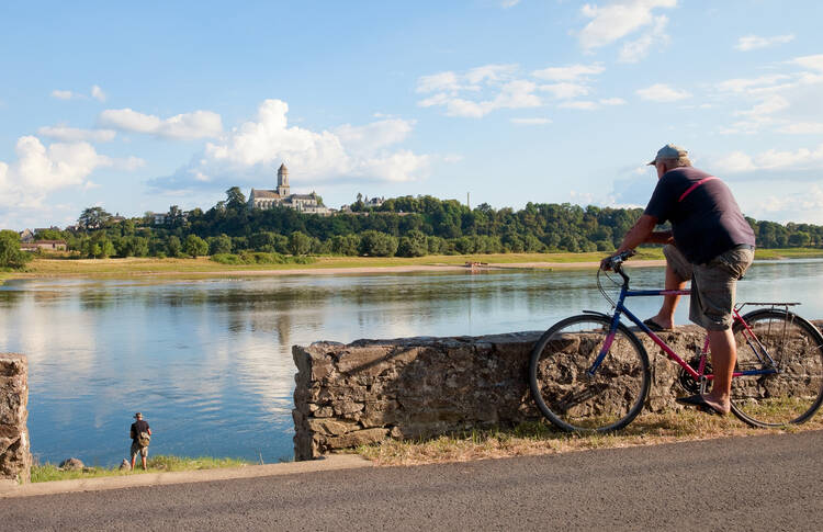 Ein Radfahrer steht an einer kniehohen Mauer und stützt seinen Fuß auf dieser ab, Blick auf den Fluss. Am Flußufer angelt ein Mann. Auf der anderen Seite des Flußes ist ein von Bäumen umgebenes Schloss und ein Dorf zu sehen.