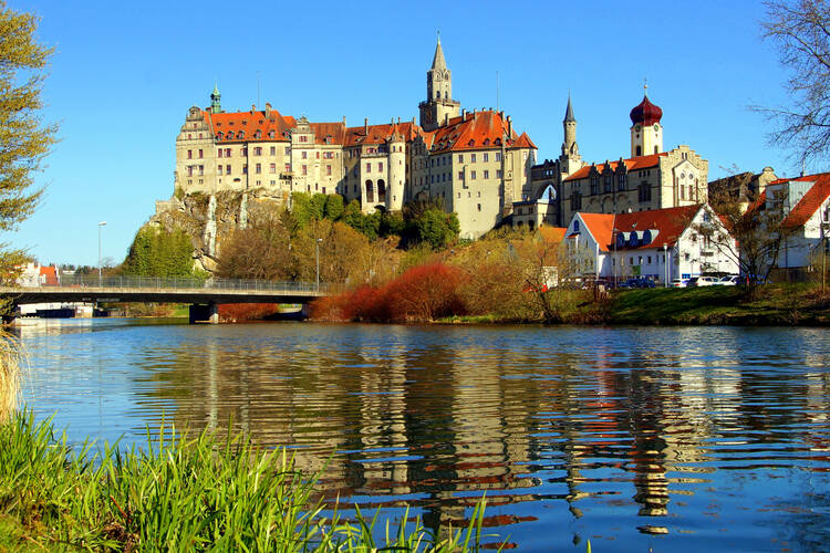 Es ist ein Fluss zu sehen. Im Vordergrund ist das grüne Ufer erkennbar. Auf der anderen Seite des Flusses ist ein Schloss, das Schloss Sigmaringen. Daneben sind kleine Häuser, ein paar Bäume. Eine Brücke führt über den Fluss in Richtung Schloss. Auf dem Fluss, die Donau, spiegelt sich das Schloss.