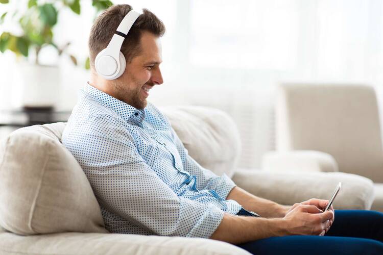 Mann mit Over Ear Kopfhörern sitzt lächelnd auf Sofa und schaut lächelnd auf Smartphone