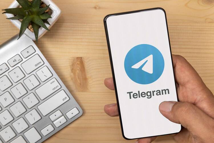 Handy mit Telegram Symbol wird an Schreibtisch in Hand gehalten