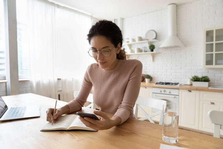 Frau sitzt an Küchentisch, schreibt auf Papier und hält das Handy in anderer Hand