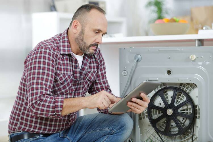 Mann kniet mit einem Tablet in der Hand vor der Rückseite einer Waschmaschine