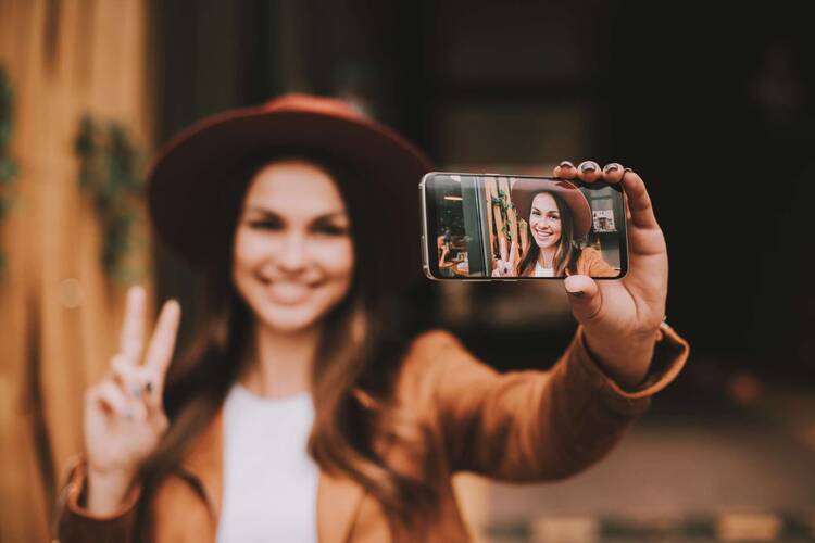 Frau im Blogger-Outfit richtet die Smartphone Kamera auf sich und macht Peace-Zeichen