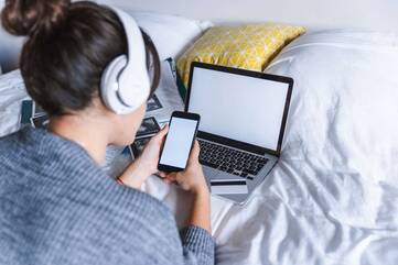 Frau mit Kopfhörern bedient Smartphone vor einem Laptop auf dem Bett