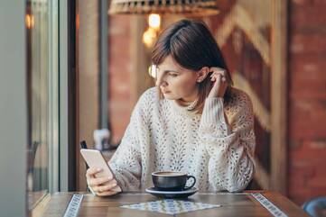 Frau sitzt auf ihr Smartphone schauend vor einer Tasse Kaffee