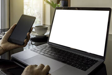 Person sitzt hält schwarzes Smartphone vor ein silbernes Notebook