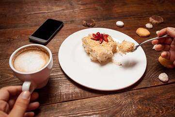 Schwarzes Handy liegt auf dem Tisch, dazu Kaffee und Kuchen auf einem Teller