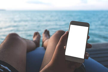 Handy in einer linken Hand, Beine und Meer im Hintergrund