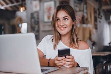 Frau sitzt lächelnd in Café vor aufgeklapptem Laptop mit Smartphone in den Händen