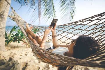 Frau liegt mit Smartphone in den Händen in einer Hängematte am Strand