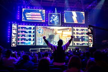 Übersicht über die am Turnier teilnehmenden Gamer auf Bildschirmen vor einem jubelnden Publikum
