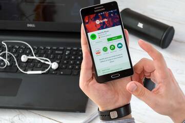 Person hält schwarzes Smartphone, welches den Google Play Store zeigt. Im Hintergund befindet sich ein Laptop, darauf liegen Kopfhörer und rechts davon eine Musikbox.