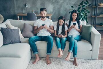 Eltern sitzen mit ihrer Tochter auf dem Sofa, alle schauen auf ihre Smartphones
