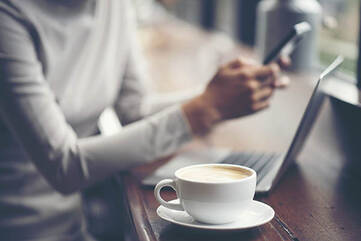 Person sitzt vor einem Tisch mit Laptop und Kaffee mit Smartphone in der Hand