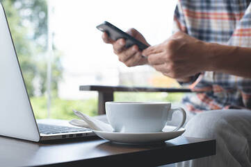Tisch mit Laptop und Tasse wo ein Mensch ein Smartphone bedient