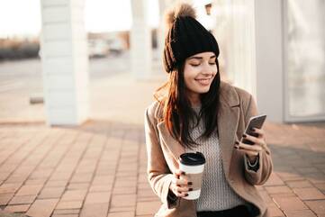 Frau sitzt draußen und hält einen Kaffee sowie ihr Smartphone in der Hand