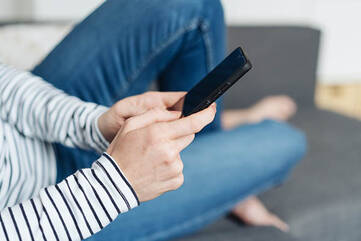 Weibliche Person hält Smartphone in beiden Händen, während sie auf einer Couch sitzt.