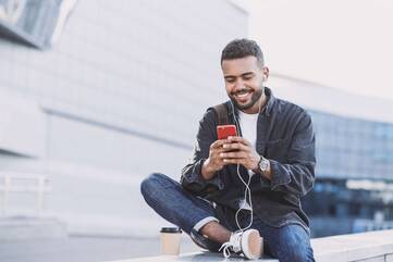 Mann sitzt mit einem Becher Kaffee und Kopfhörern in den Ohren auf einer Mauer und schaut lächelnd auf sein Handy