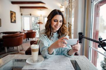 Eine Frau sitzt in einem Kaffee und lächelt ihr Smartphone an, welches sich in ihren Händen befindet.