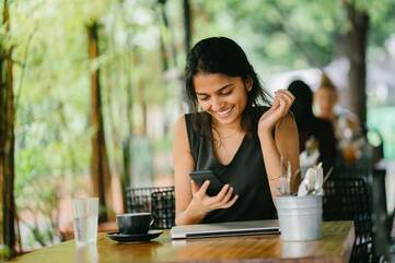 Junge Frau sitzt an einem Tisch und lächelt ihr schwarzes Smartphone in ihrer Hand an.