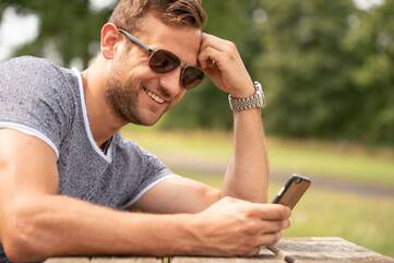Mann sitzt mit Sonnenbrille im Park und halt ein schwarzes Smartphone in der Hand.