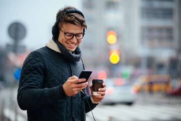 Mann mit Winterjacke, Kopfhörern und Coffee-to-go bedient sein Smartphone