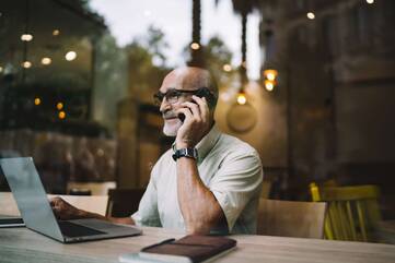 Älterer Mann in Café sitzt vor Laptop und Notizbuch und telefoniert