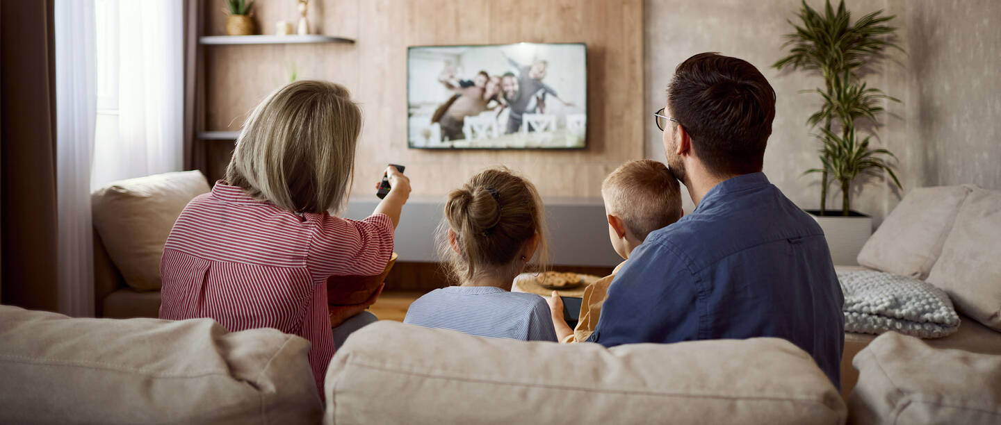 Man sieht eine Familie von hinten, die auf einem Sofa sitzt und gemeinsam TV schaut