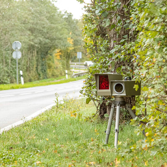 Ein mobiler Blitzer steht in einer Hecke an einer Landstraße.