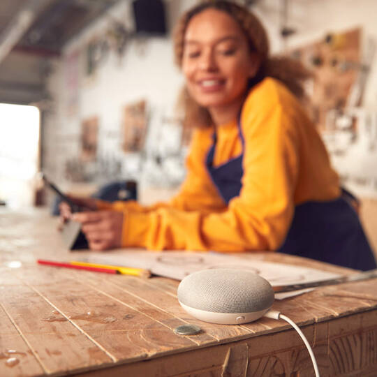 Eine Frau arbeitet am Tablet und blickt auf ihren Smart Speaker.