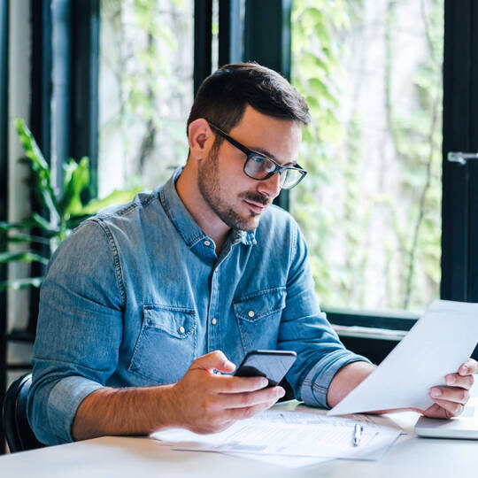 Ein Mann schaut aufs Handy und liest parallel etwas von einem Zettel ab.