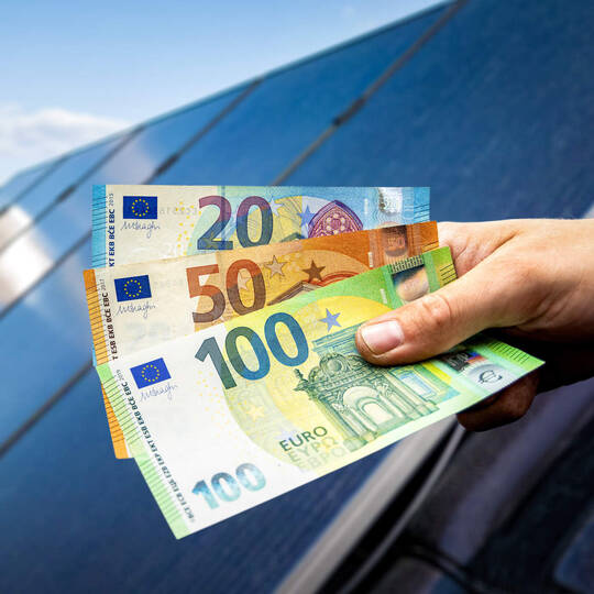 Ein Person hält ein paar Geldscheine in der Hand. Im Hintergrund ist eine Solaranlage zu sehen.