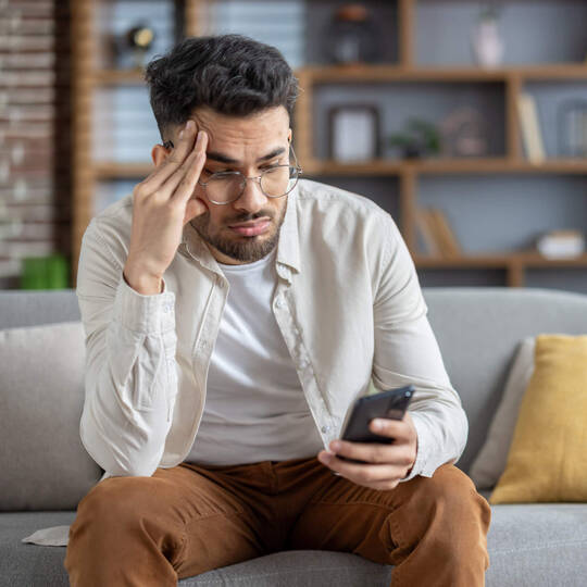 Ein Mann sitzt auf dem Sofa und blickt ratlos auf sein Handy.