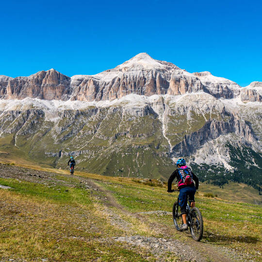 Mountainbiker fahren über eine Wiese in Richtung des Sellastocks der Dolomiten