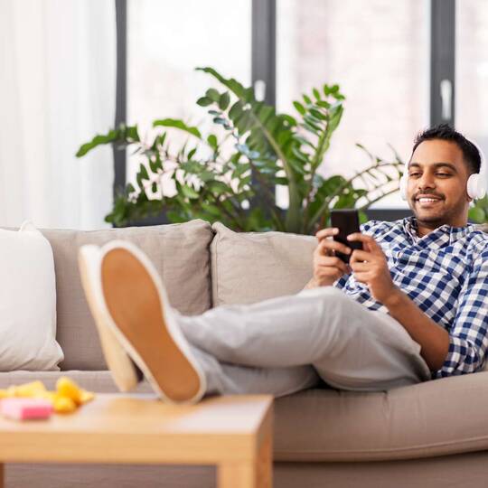 Mann sitzt lächelnd mit Kopfhörern auf dem Sofa und schaut auf sein Smartphone