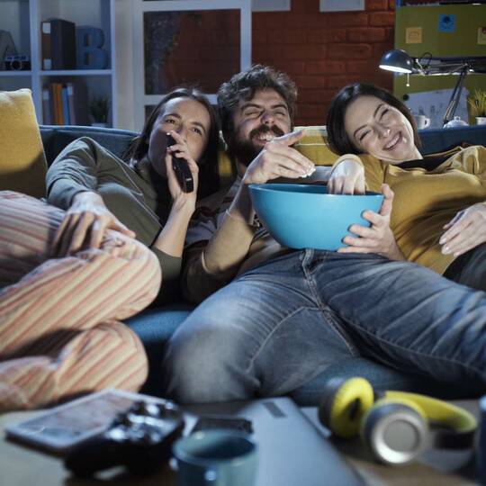 Drei Personen sitzen auf Couch und schauen auf Fernseher