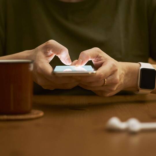 Zwei Hände bedienen iPhone, Apple Watch an Handgelenk, AirPods liegen davor, Kaffeetasse steht auf dem Tisch