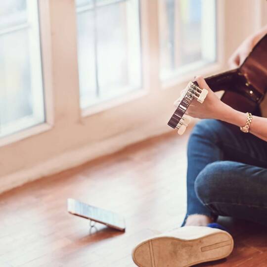 Mensch sitzt im Schneidersitz mit Gitarre auf dem Schoß auf dem Boden mit aufgestelltem Smartphone vor sich