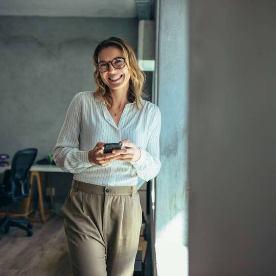 eine Frau steht im Büro und schaut lächelnd voon ihrem Smartphone auf