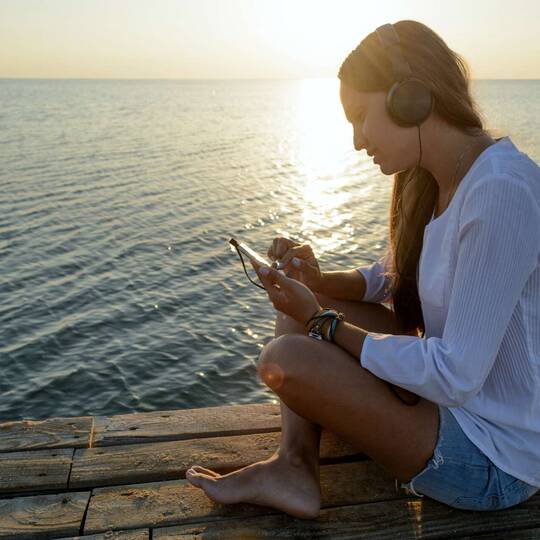 Frau sitzt im Schneidersitz auf Steg am Wasser mit Over Ear Kopfhörern und schaut auf Smartphone in ihren Händen