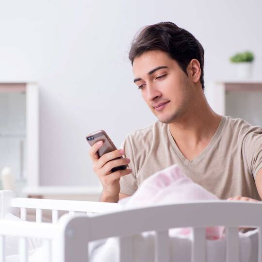 Mann an Babybett schaut auf ein Smartphone